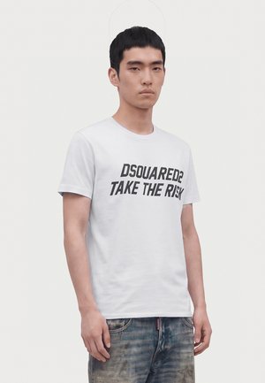 Млад мъж с къса черна коса носи бяла тениска с текст "DSQUARED2 TAKE THE RISK" и износени сини дънки на едноцветен светъл фон.