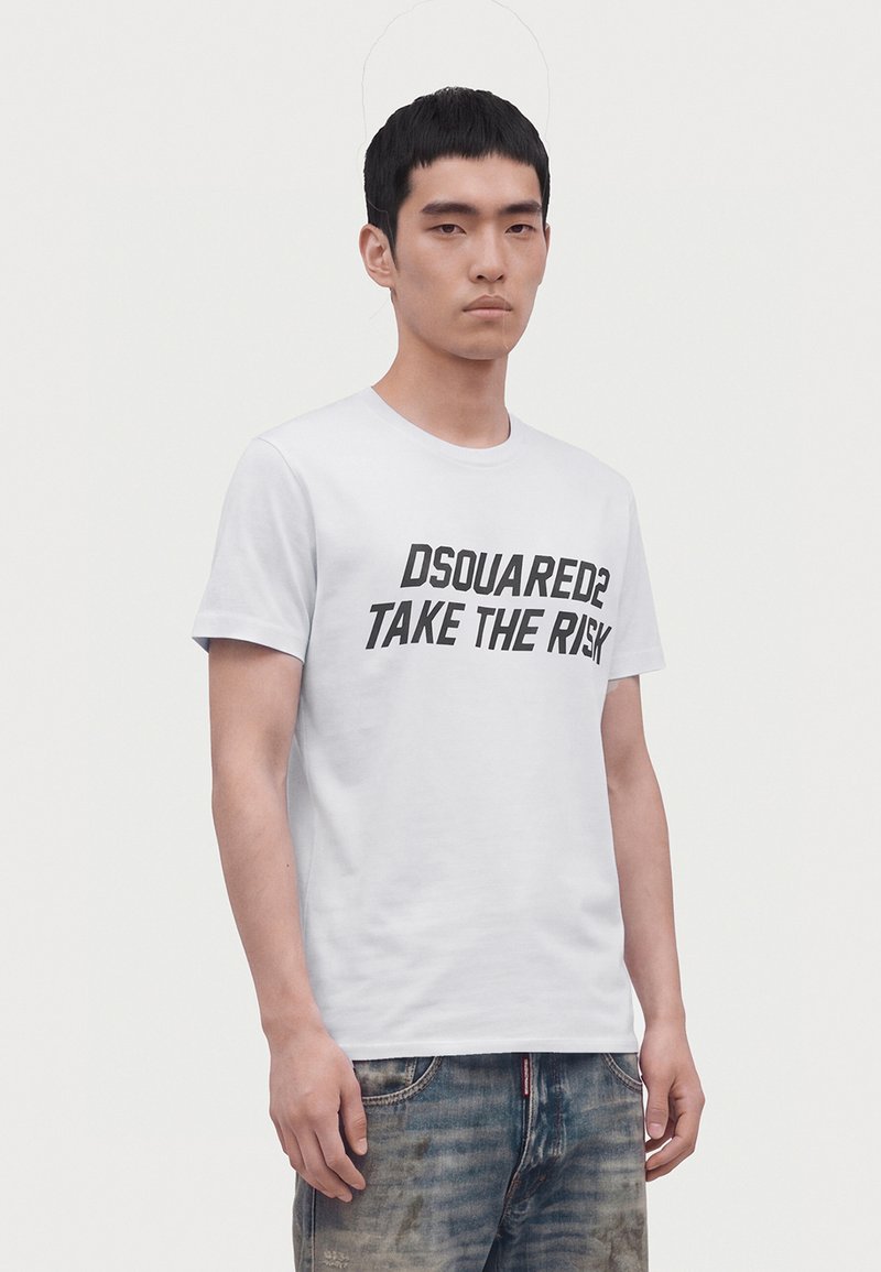 Jeune homme aux cheveux courts noirs porte un T-shirt blanc avec le texte "DSQUARED2 TAKE THE RISK" et un jean bleu délavé sur un fond clair uni.