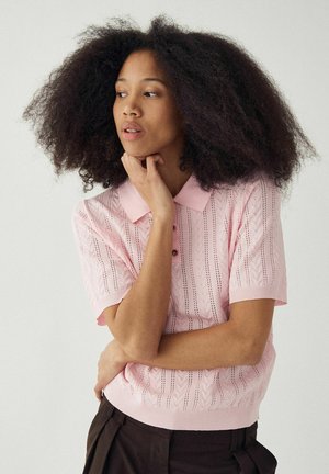 Kvinne med voluminøst krøllete hår, iført en rosa mønstret genser med korte ermer og mørke bukser, hviler haken på hånden og ser sidelengs.