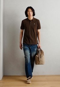 Braunes Polohemd, blaue Jeans, beiger Sneaker, trägt eine beige Tasche. Gerade geschnittene Jeans mit verblasstem Finish, das Polohemd hat ein kleines Logo.