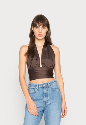 Νεαρή γυναίκα με μακριά καστανά μαλλιά, φορώντας χρυσά σκουλαρίκια, σκούρο καφέ τοπ χωρίς μανίκια και ψηλόμεσα ανοιχτόχρωμα τζιν, που στέκεται μπροστά σε λευκό φόντο.