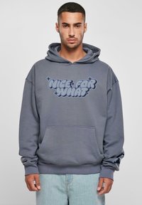 Grå oversized hettegenser med en stor blå grafisk tekst "NICE FOR WHAT" på forsiden, kengurulomme og ribbestrikkede ermer.