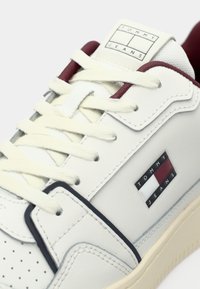 Sapatilha de pele branca com detalhes texturizados, atacadores creme e um detalhe em azul marinho escuro. Apresenta o logótipo "TOMMY JEANS" na lateral.