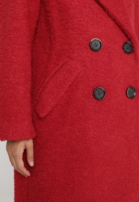 Rød dobbeltradet frakke i uldblanding med tekstureret stof, store sorte knapper og sidelommer, med bred krave og afslappet pasform.
