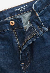 Jeans din denim skinny fit de culoare albastru închis, cu fermoar la bază, nasture de cupru și cusături bej pe margini. Mărimea 34 R.