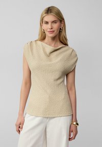 Beige textuur top met een gedrapeerde halslijn en kapmouwtjes, gemaakt van een zachte stof; gecombineerd met wijde witte broeken voor een relaxte pasvorm.