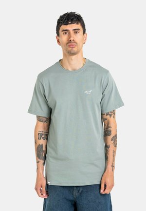Hombre con cabello corto y oscuro y tatuajes en ambos brazos lleva una camiseta verde claro y jeans azules sobre un fondo claro y uniforme.