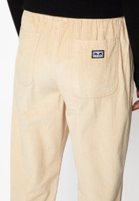 Beige kordflöjtsbyxor med vertikala ribbor, elastisk midja och två bakfickor med en liten logomärke på högra fickan.