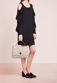 Robe noire avec épaules dénudées et manches à volants, associée à un sac à main gris clair portant un logo. Le modèle porte des mocassins noirs brillants.