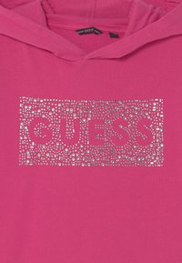 Sudadera rosa con un logo "GUESS" plateado y tachonado, con patrones geométricos y acentos brillantes en la parte delantera. Textura de tela suave.