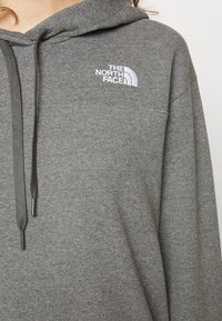 Gros plan sur un sweat à capuche gris avec cordons de serrage et logo blanc "The North Face" brodé sur la poitrine.