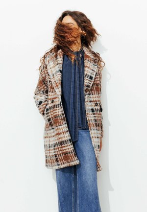 Manteau à carreaux présentant des couleurs marron, blanc et noir ; tissu à texture douce ; design croisé avec de grands boutons ; poches latérales.