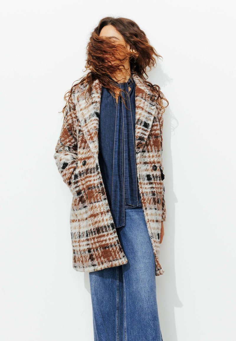 Manteau à carreaux présentant des couleurs marron, blanc et noir ; tissu à texture douce ; design croisé avec de grands boutons ; poches latérales.