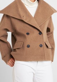 Brun ullblandad dubbelknäppt kappa med bred krage, svarta knappar och två framfickor, bärs över en vit polotröja.