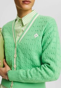 Grüne Strickjacke mit strukturiertem Muster, V-Ausschnitt und cremefarbenem Besatz. Verfügt über Knöpfe vorne und ein kleines Emblem auf der Brust.