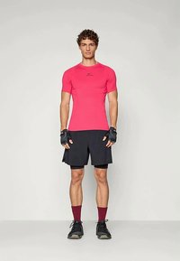 Mann står iført en lys rosa idrettsgenser, svarte shorts, svarte fingerløse hansker, maroon sokker og svarte løpetrenere mot en enkel bakgrunn.