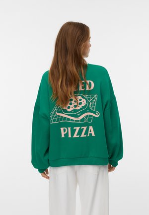 Donna con lunghi capelli castani che indossa una felpa verde con una grafica di pizza rosa e il testo "Baked Pizza" sulla schiena, abbinata a pantaloni bianchi.