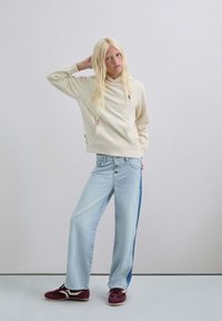 Mujer rubia con sudadera color crema, jeans de pata ancha azul claro con franja lateral oscura, y zapatillas burdeos, de pie contra un fondo claro y liso.