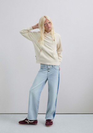 Mujer rubia con sudadera color crema, jeans de pata ancha azul claro con franja lateral oscura, y zapatillas burdeos, de pie contra un fondo claro y liso.