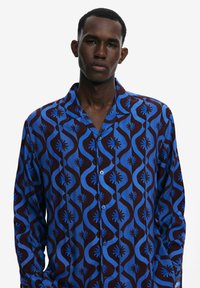 Blue and brown patterned button-up shirt with a fold-down collar, featuring wave-like designs and starburst motifs on a lightweight fabric.