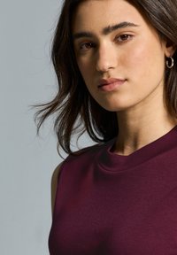 Top sin mangas burdeos con escote redondo, hecho de un tejido suave. El modelo presenta cabello largo y oscuro, con un maquillaje minimalista y un pendiente sutil.