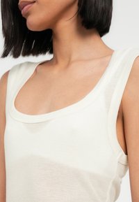 Ριμπ τοπ αμάνικο σε απαλό κρεμ χρώμα, με στρογγυλή λαιμόκοψη και φαρδιές τιράντες. Το υλικό φαίνεται ελαφρύ και εφαρμοστό.