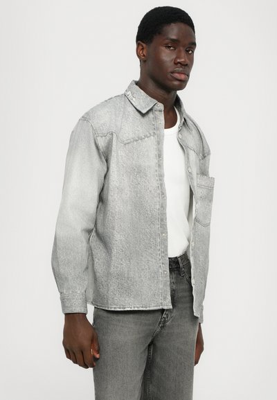 Camasa din denim gri cu butoni pe față, guler ascuțit și buzunar pe piept. Materialul are o textură subtilă în diagonală. Purtați peste un tricou alb.