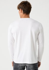 Camisa blanca de manga larga con cuello redondo y textura suave, con costuras mínimas y dobladillo trasero recto. Combinada con pantalones grises.