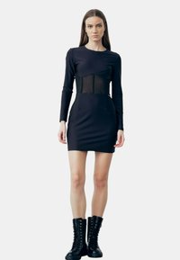 Zwart langemouwen jurk met een aansluitend lijfje, een doorzichtig netpaneel op de taille en zijzakken. Gecombineerd met zwarte veterschoenen.