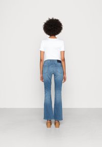 Jean flare bleu avec une taille ajustée et des poches arrière, associé à une chemise blanche à manches courtes. Le modèle se tient de dos sur un fond neutre.
