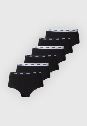 Femme debout pieds nus, portant un soutien-gorge noir et un shorty garçon noir de marque sur un fond clair uni.