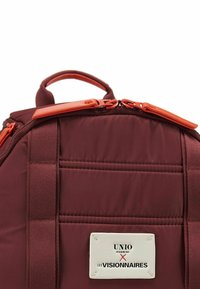 Mochila color burdeos con textura acolchada, acentos en naranja de cremallera y detalles cosidos. Presenta una etiqueta con la marca "UNIO" y "LES VISIONNAIRES".