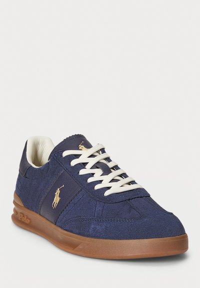 Navy blue suede sneaker with white laces, brown rubber sole, and gold polo player logo on side and tongue.