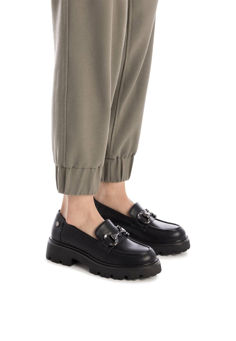 Mocasines de cuero negro con suela gruesa y detalle de cadena plateada. Combinados con pantalones verde oliva con dobladillo, resaltando el diseño del calzado.