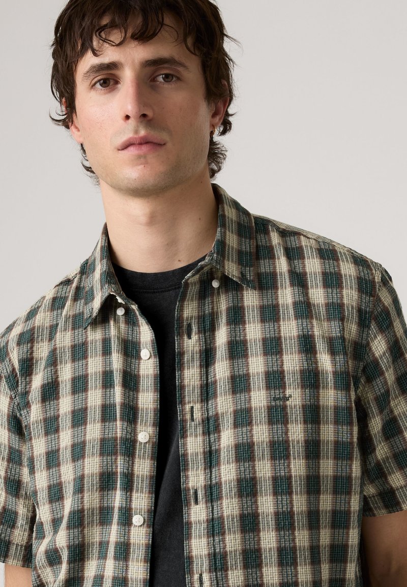 Joven con cabello oscuro vestido con una camisa de cuadros verde y beige abotonada sobre una camiseta negra, de pie frente a un fondo claro y liso.