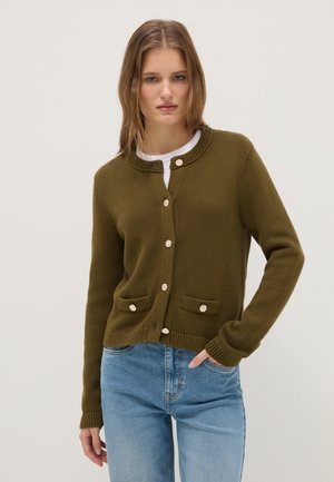 Junge Frau trägt eine olivgrüne, geknöpfte Strickjacke mit Taschen über einem weißen Top und hellblauen Jeans, steht vor einem schlichten Hintergrund.