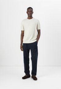 Licht beige kortetermijntrui met korte mouwen, ronde hals, aansluitend bij de taille, gecombineerd met marineblauwe op maat gemaakte broeken en bruine schoenen. Gelaagd oppervlak.