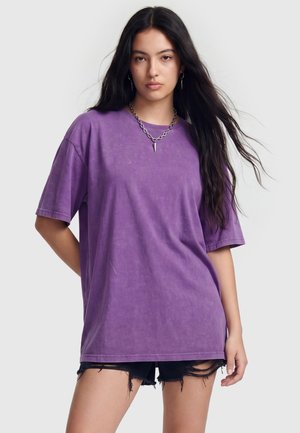 Ung kvinde med langt mørkt hår iført en lilla oversized t-shirt, sorte hullede shorts, et kædehalskæde og øreringe, poserer med den ene hånd bag ryggen.