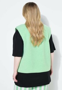 Fleeceväst i mintgrön med en texturerad yta, kombinerad med en svart T-shirt. Randiga byxor fulländar outfiten. Ingen uppenbar hårdvara visas.