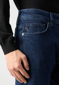 Temno modri kavbojke z rahlo zbledelim izgledom, s majhno oranžno oznako "BOSS" na pasu in klasičnim krojem.