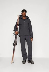 Muž ve černém lyžařském oblečení držící lyže vzpřímeně, oblečený v lyžařských botách a brýlích, stojící na jednobarevném pozadí.