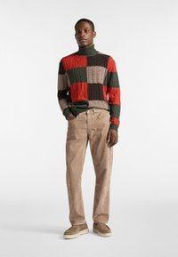 Rot gestrickter Rollkragensweicher mit Blockmustern in Rot, Grün, Braun und Schwarz; kombiniert mit braunen Cordhosen; das Modell steht mit den Händen in den Taschen.