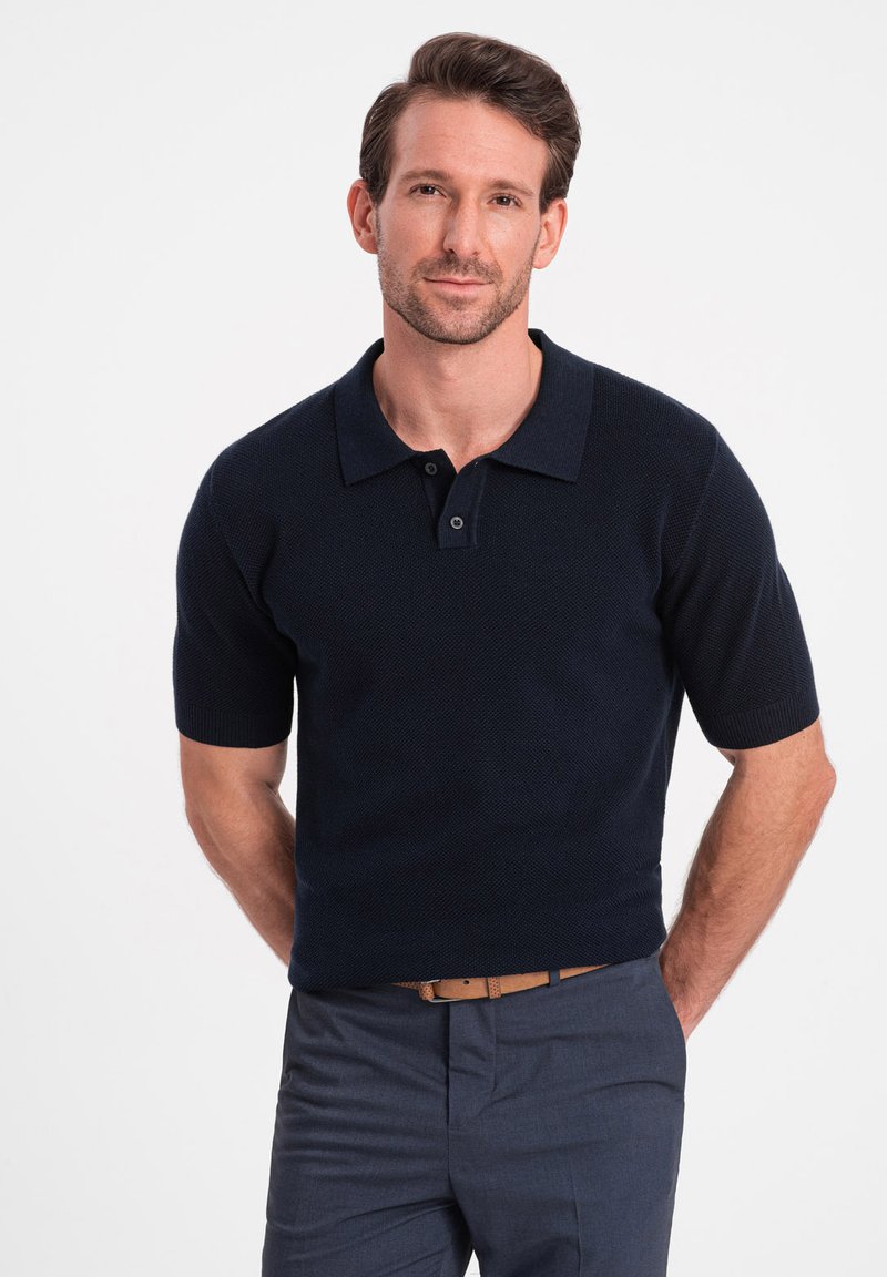 Polo à manches courtes en navy, en tissu tricoté texturé. Présente un col, deux boutons et une coupe ajustée. Associé à un pantalon gris ajusté.