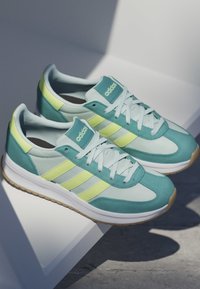 Paar türkisfarbene und hellgelbe Adidas-Sneaker mit weißen Schnürsenkeln und Gummisohlen, die auf einer weißen Oberfläche mit schattigem Hintergrund liegen.