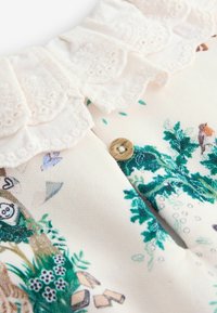 Puuvillavaate, jossa on aaltoileva pitsikaulus, puunappi sekä värikkäitä kukka- ja eläinkuoseja kermanvärisellä pohjalla.