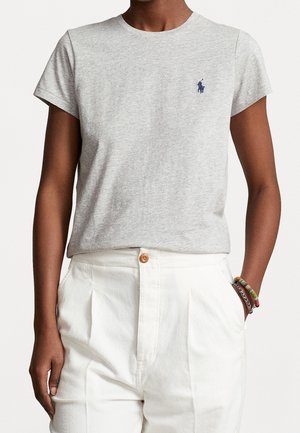 Persona con una camiseta de manga corta color gris jaspeado con un pequeño logo azul oscuro y pantalones blancos de talle alto con pliegues, con una mano en el bolsillo.