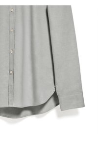 Ljusgrå skjorta med knappar och en slät yta samt rundad nederkant. Har beiga knappar och en enkel manschett på den långa ärmen.