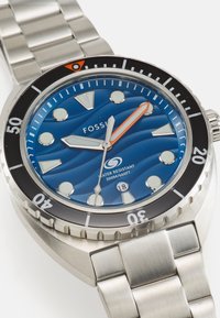Orologio Fossil con cinturino in acciaio inossidabile, quadrante con motivo a onde blu, indicatori bianchi, lancetta dei secondi arancione e lunetta girevole nera. Impermeabile fino a 200 m.