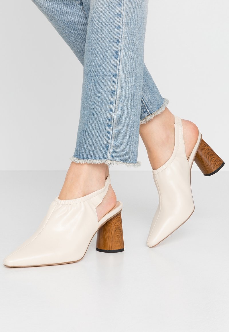 Pieds portant des escarpins beige clair à bride arrière avec talons blocs en bois et jeans bleu clair raccourcis à ourlets effilochés.
