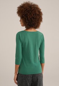 Grön, figurnära långärmad topp med rundad halsringning och trekvartslånga ärmar. Slät yta, enkel design, tillsammans med texturerade grå byxor.